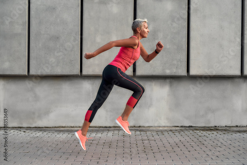 Full of energy. Side view of active middle aged woman in sport clothing jumping while exercising outdoors photo