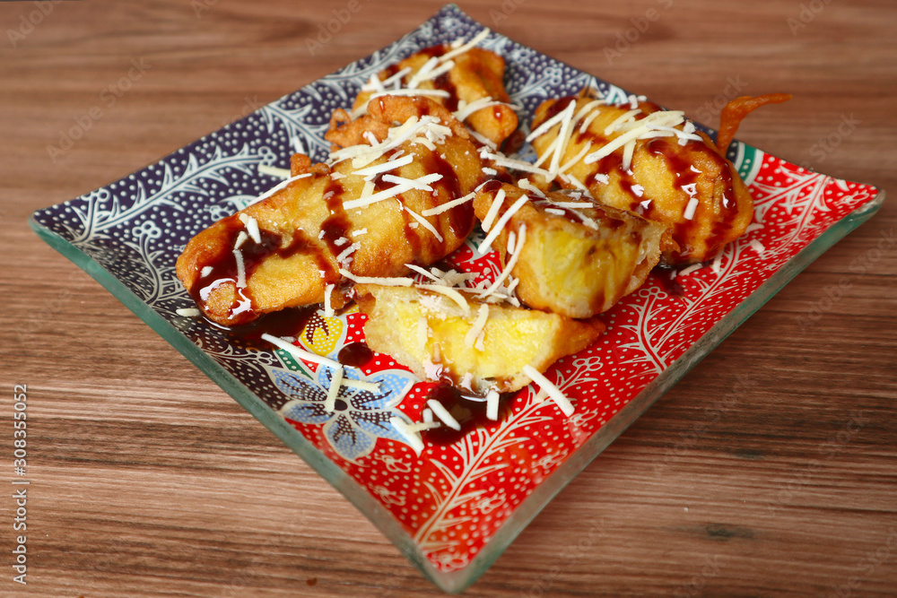 Fried banana or pisang goreng with chocolate sweetened condensed milk and cheese on batik plate with wood background Fried banana or pisang goreng with chocolate sweetened condensed milk and cheese on batik plate with wood background