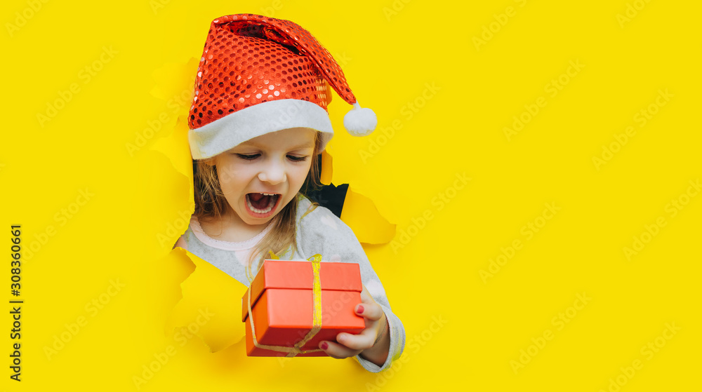 A cute little surprised child in a red Santa hat looks out of a hole in yellow paper and holds a box with a gift in her hand. Christmas and New Year concept. Bright background, copy space. A cute little surprised child in a red Santa hat looks out of a hole in yellow paper and holds a box with a gift in her hand. Christmas and New Year concept. Bright background, copy space.