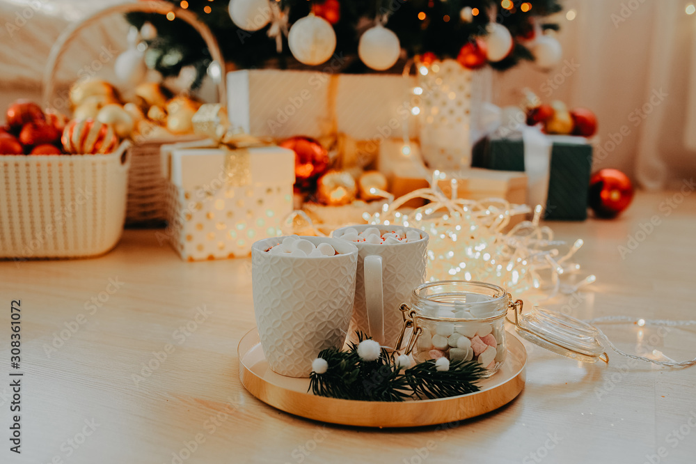 two mugs with a drink and marshmallows, on a gold tray with a can of marshmallows and a sprig of a Christmas tree, against the background of a Christmas tree in the lights and gifts under it