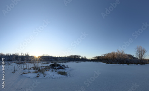 Scenic calm winter view of a frozen snow covered beaver pond with a sun star as the sun descends into the trees.