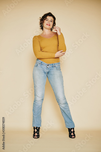 Pretty smiling young woman in jeans and mustard top with long sleeves posing at beige wall