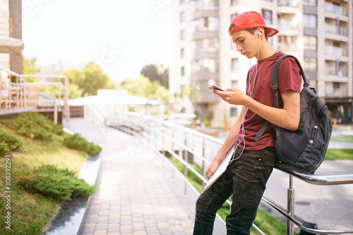 Young man  stands with backpack and holds smartphone, in the city.   Teenage boy is using mobile phone, outdoors.  Caucasian teenager in casual clothes with cell phone, urban scene.