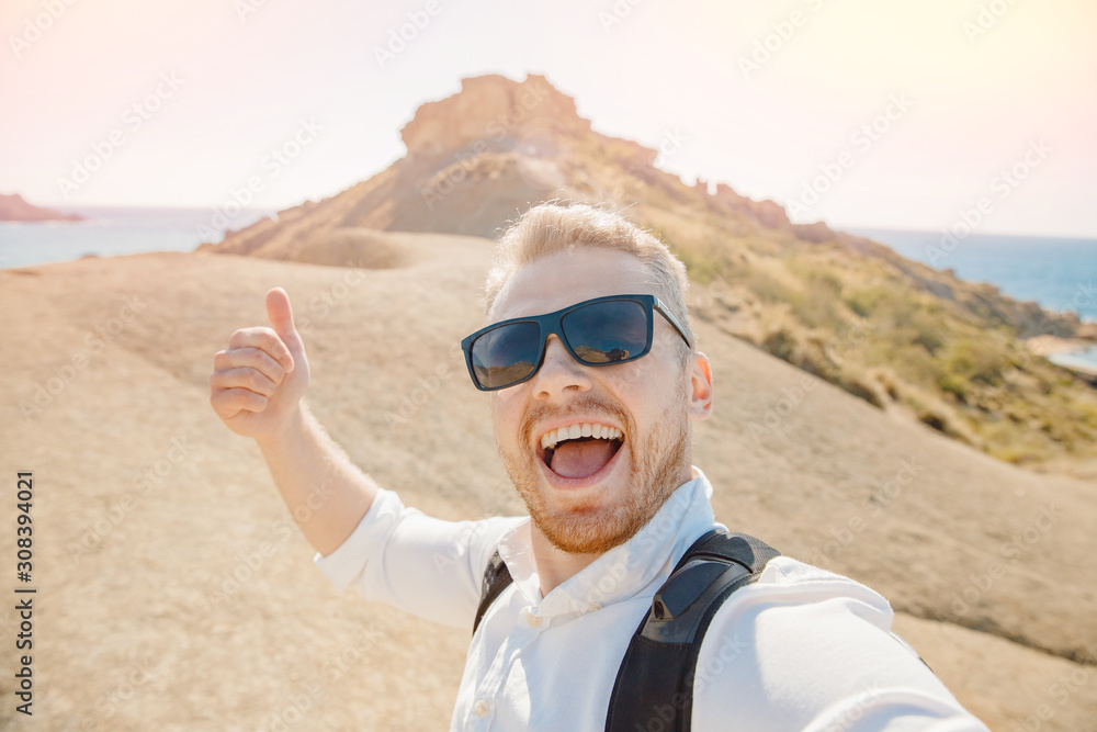 Male traveler makes selfie photo on background of blue sea, sand beach in sunglasses and with backpack. Travel concept. Gold bay Malta