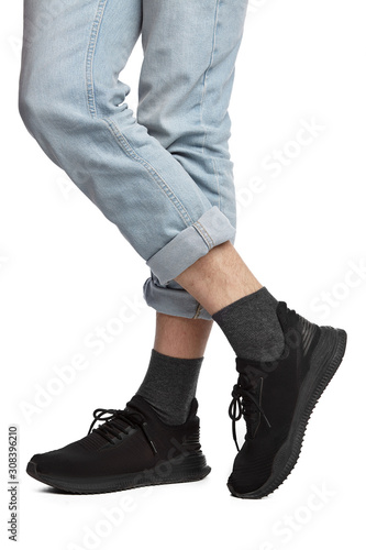 Cropped shot of a man's cross-legs in blue jeans and black sneakers, staying on a white background. It is black socks on his foots. 