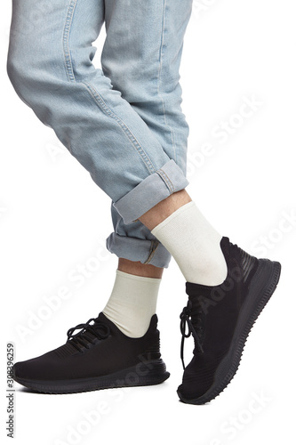 Cropped shot of a man's cross-legs in blue jeans and black sneakers, staying on a white background. It is white socks on his foots. 