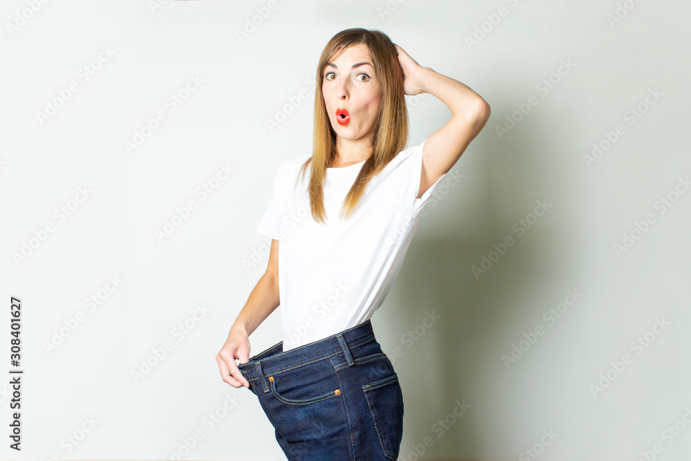 Young woman with a surprised face in oversize jeans on a light background. The concept of shock from a diet, a good result from losing weight, a good diet, fitness Young woman with a surprised face in oversize jeans on a light background. The concept of shock from a diet, a good result from losing weight, a good diet, fitness