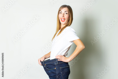 Young woman with a surprised face in oversize jeans on a light background. The concept of diet, a good result, weight loss, fitness