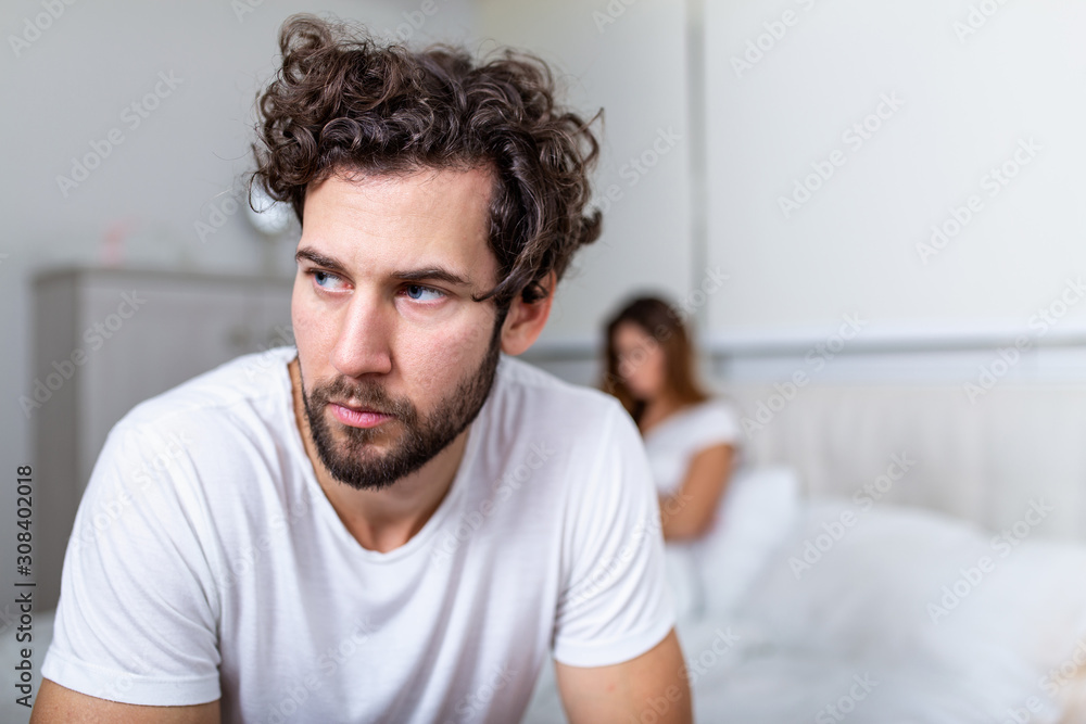 Sad thoughtful man after arguing with girlfriend.Relationship difficulties, conflict and family concept,unhappy couple having problems at bedroom. Sad guy sitting on bed,girlfriend in the background - obrazy, fototapety, plakaty 