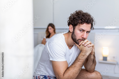 Sad thoughtful man after arguing with girlfriend.Relationship difficulties, conflict and family concept,unhappy couple having problems at bedroom. Sad guy sitting on bed,girlfriend in the background