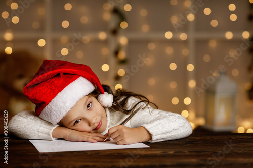 Happy little child girl in santa hat writing wish list or letter to Santa Claus on background with lights. Merry Christmas
