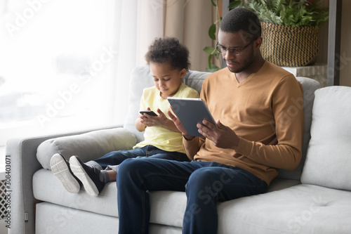Black father and little son use gadgets sitting on couch