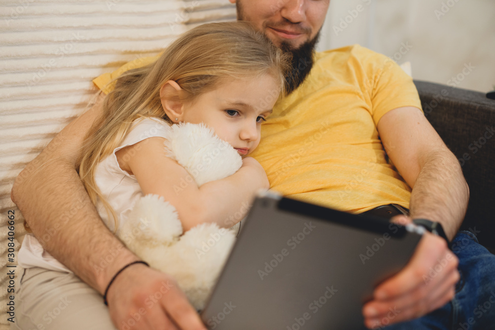 Close up portrait of a cute little girl leaning on her father chest embracing a white teddy bear and looking to her fathers tablet. Close up portrait of a cute little girl leaning on her father chest embracing a white teddy bear and looking to her fathers tablet.
