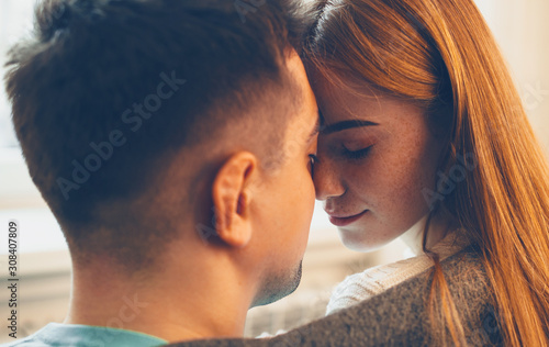 Close up side view portrait of a beautiful woman with red hair and freckles and her man sitting face to face with eyes closed before kissing at home.