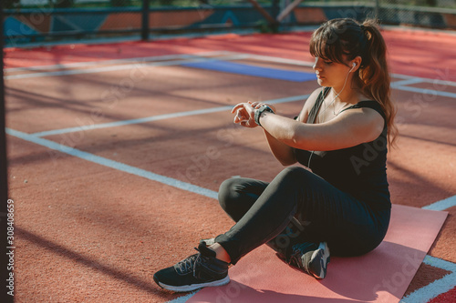 Portrait of a lovely plus size woman sitting on the ground in a sport park looking at her sport watch before doing losing weight exercises.