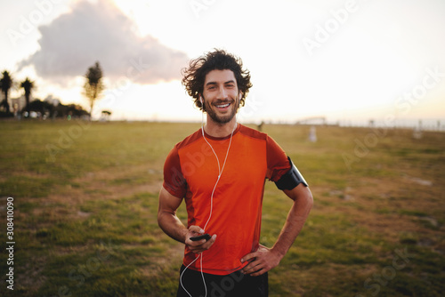 Portrait of a happy fitness young man in earphones holding mobile phone while standing in park looking at camera