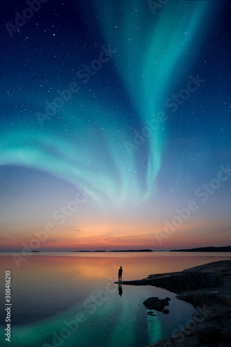 A man standing by a calm water and looking at the northern lights on the sky with reflections from the water