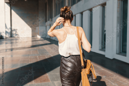 Refined young woman in leather skirt and silk blouse walking confident near a building.