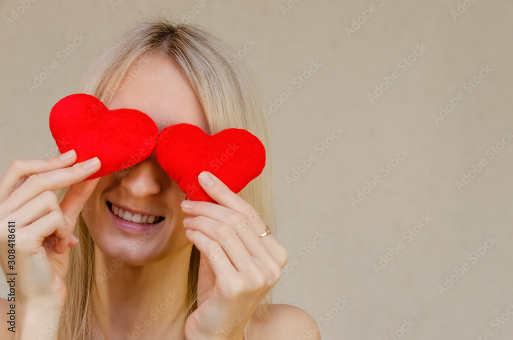 Lifestyle concept: attractive blonde woman with a radiant smile, having two small red hearts in her hands, closing her eyes with heart symbols, standing on a light background. Valentine's day Lifestyle concept: attractive blonde woman with a radiant smile, having two small red hearts in her hands, closing her eyes with heart symbols, standing on a light background. Valentine's day
