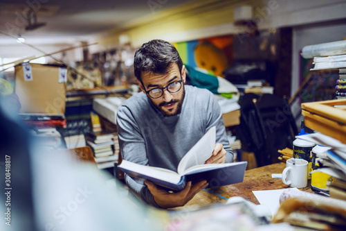 Young attractive caucasian bearded serious man with eyeglasses sitting in back of bookstore and reading interesting book.