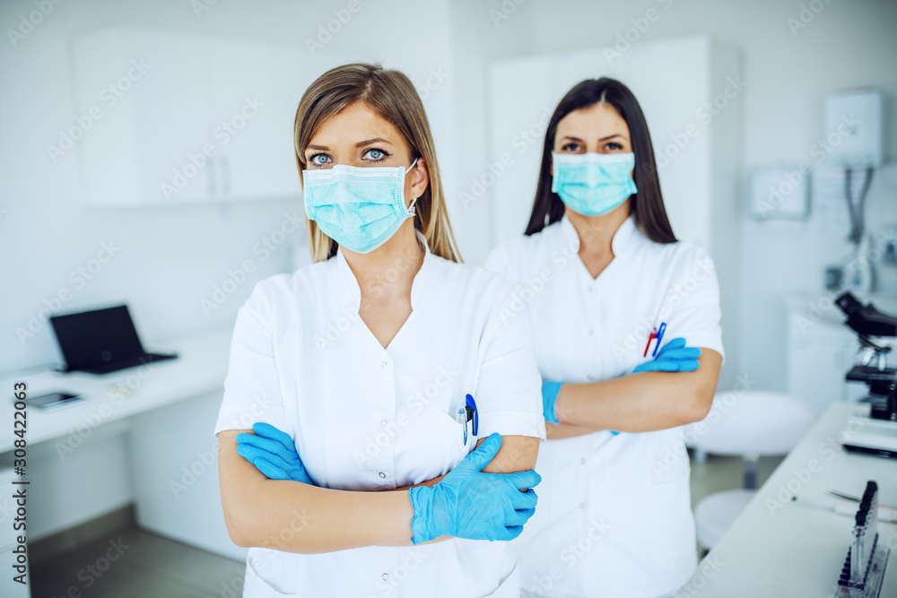 Blonde female lab assistant in white uniform, with rubber gloves and mask standing with arms crossed. In background is her colleague. Blonde female lab assistant in white uniform, with rubber gloves and mask standing with arms crossed. In background is her colleague.