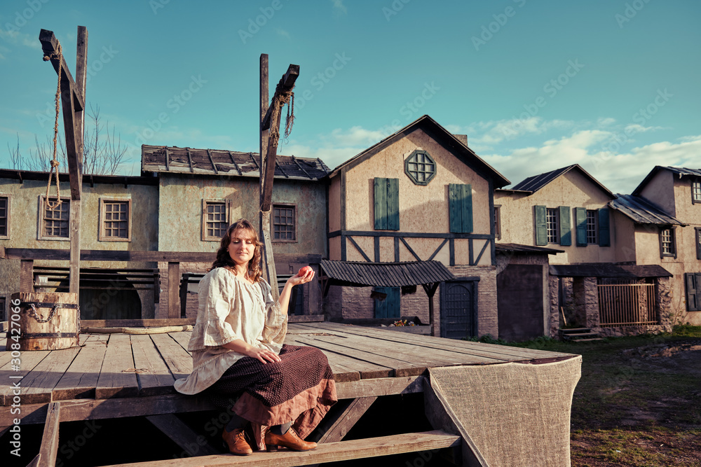 A woman with an apple sits on the steps of a wooden platform with gallows. Sunset on the background of old houses at the execution site A woman with an apple sits on the steps of a wooden platform with gallows. Sunset on the background of old houses at the execution site