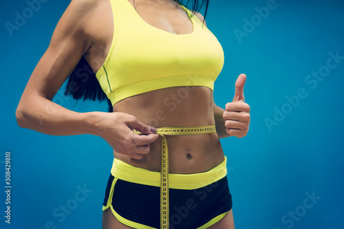 Crop shot of fitness woman in sport outfit measuring waistline with metric tape and showing thumb up. Good results of exercising in gym. Perfect shape of sportswoman. Achievments in body perfection. photo