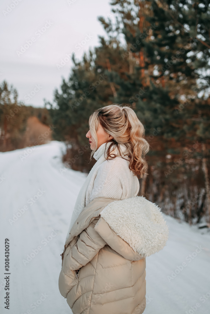 Happy attractive beautiful lonely blonde in a white sweater, warm jacket and jeans walks, enjoys freedom and tranquility on a snowy road in the forest in winter.