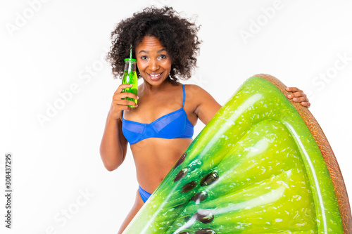 attractive african girl in a blue separate swimsuit holds a swimming mattress and drinks a cocktail on a white wall