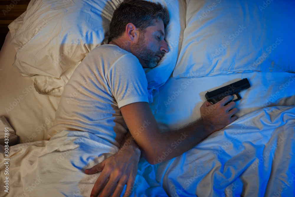 night portrait of young stressed and paranoid American man in bed sleeping and holding gun fearing intruder breaking in home in paranoia and self defense concept night portrait of young stressed and paranoid American man in bed sleeping and holding gun fearing intruder breaking in home in paranoia and self defense concept