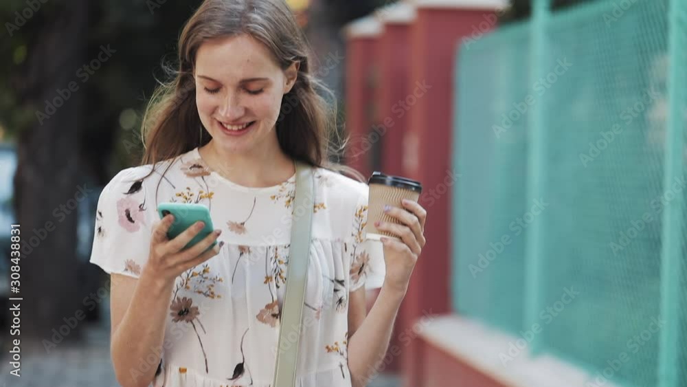 Young Beautiful Girl Wearing White Flower Dress with Bag Cross her Shoulder Holding Coffee Cup and her Smartphone and Making Voice Message or Using Voice Commander. Woman Walking at the Street. Young Beautiful Girl Wearing White Flower Dress with Bag Cross her Shoulder Holding Coffee Cup and her Smartphone and Making Voice Message or Using Voice Commander. Woman Walking at the Street.