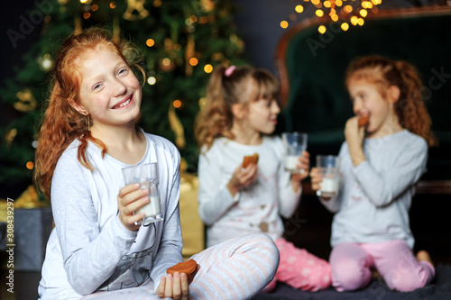 Children drink milk and eat cookies on Christmas Eve. Sisters in pajamas. The concept of Merry Christmas, holidays, family and gifts.