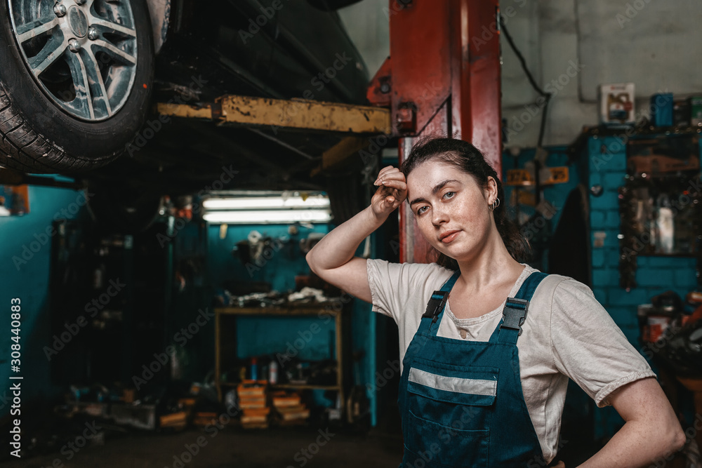 The concept of small business, feminism and women's equality. A young woman mechanic wipes sweat from her forehead, on the lift is a car