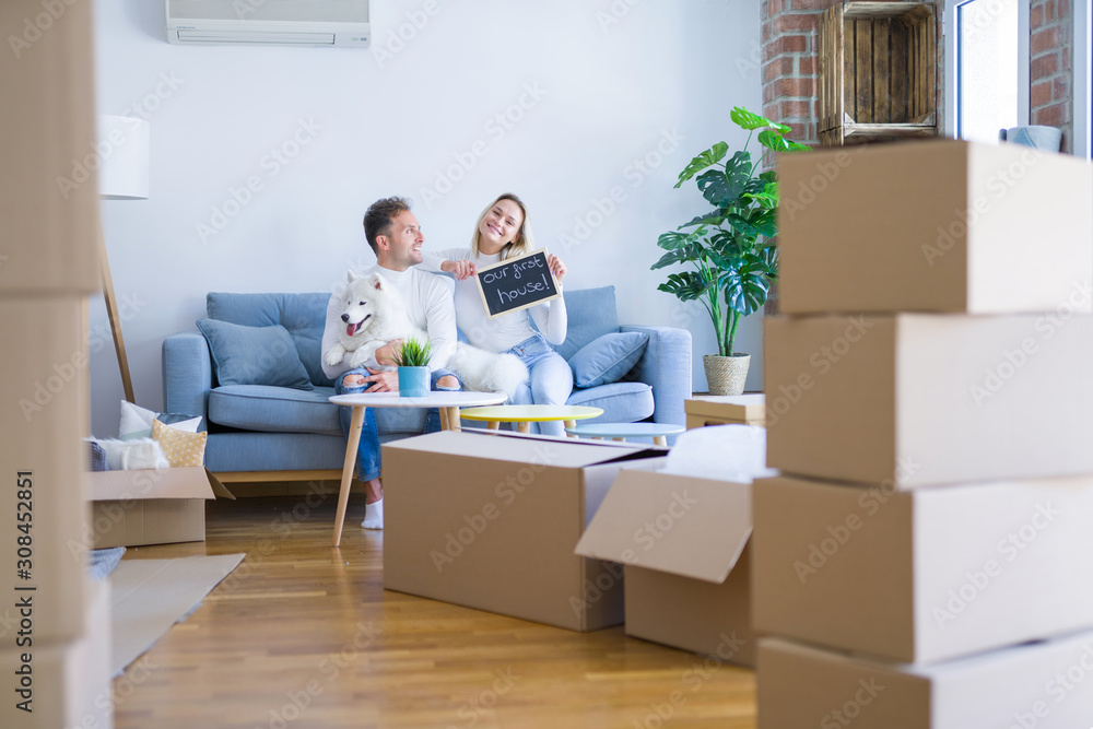 Young beautiful couple with dog sitting on the sofa holding blackboard with message at new home around cardboard boxes