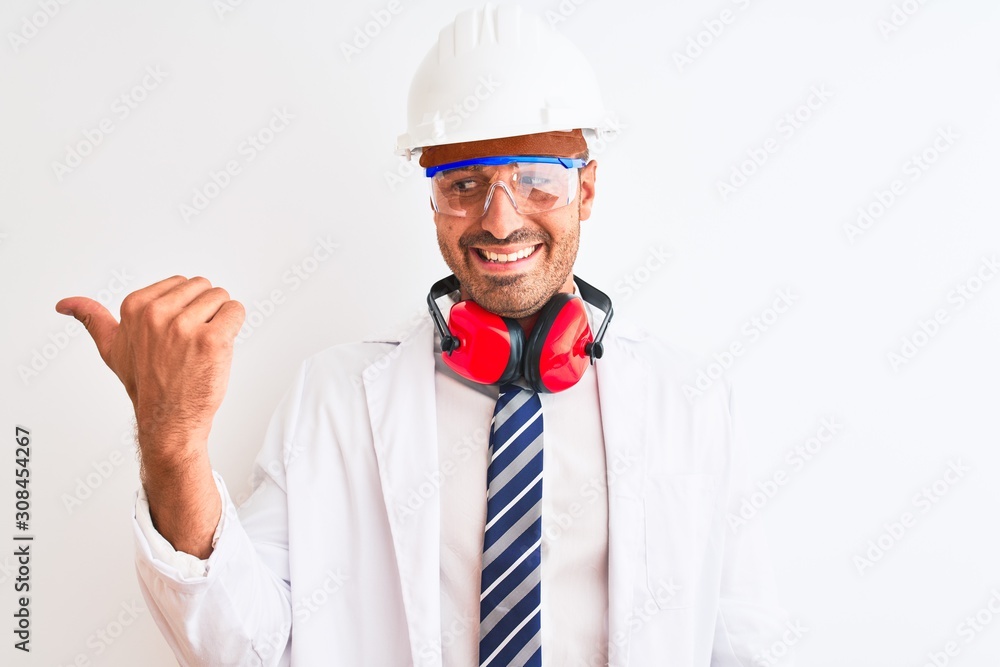 Young chemist man wearing security helmet and headphones over isolated background smiling with happy face looking and pointing to the side with thumb up. Young chemist man wearing security helmet and headphones over isolated background smiling with happy face looking and pointing to the side with thumb up.