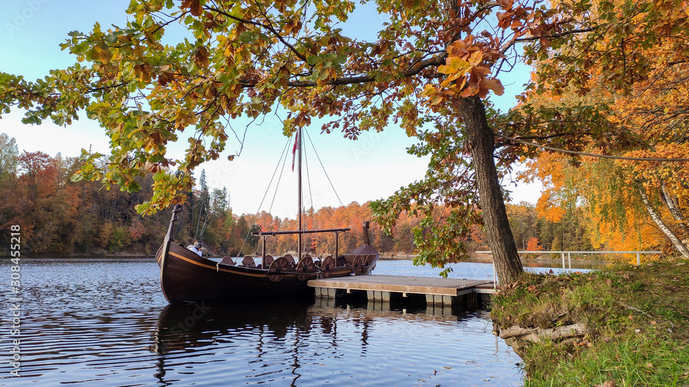 The Tourist Boat in the Style of a Viking Ship on the River Perse. Autumn Old Koknese Castle Ruins. Perse is a Tributary to Daugava River in Latvia. The Tourist Boat in the Style of a Viking Ship on the River Perse. Autumn Old Koknese Castle Ruins. Perse is a Tributary to Daugava River in Latvia.