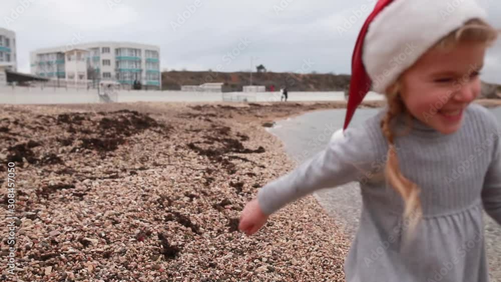 Сheerful little girl in santa hat runs along the shore Сheerful little girl in santa hat runs along the shore