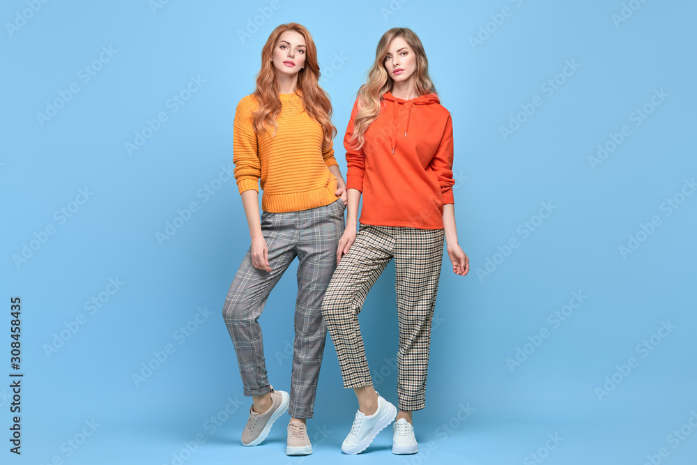 Fashionable woman in stylish outfit, makeup posing on blue. Two Beautiful blonde redhead tomboy girl, trendy orange jumper, hoody, fashion hair. Cheerful beautiful sister friend, colorful concept