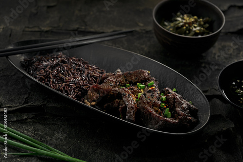 Sticky Asian beef stir fry in salty sweet sauce with black rice in a black clay plate.