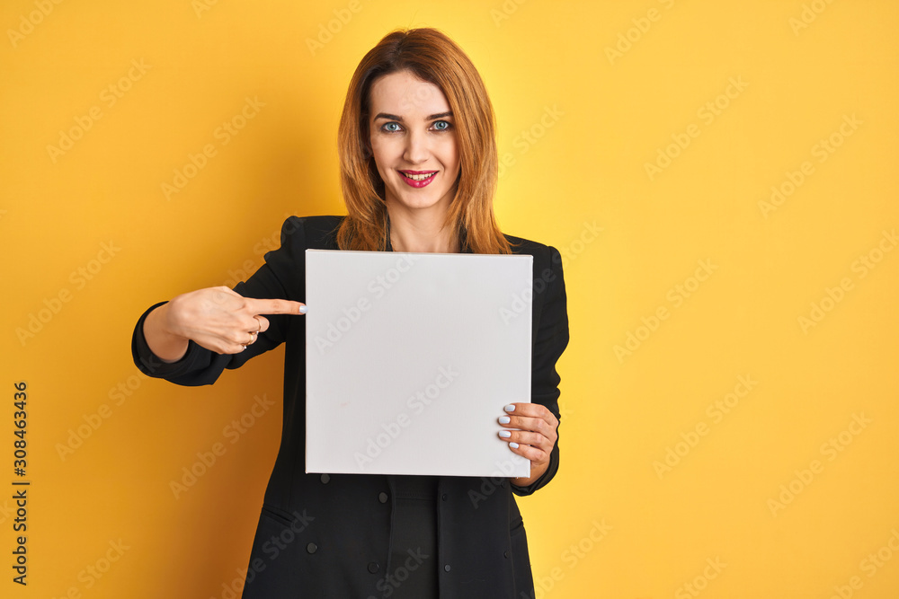 Naklejka premium Young redhead businesswoman with clear eyes holding banner over isolated yellow background very happy pointing with hand and finger