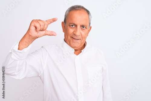 Senior grey-haired man wearing elegant shirt standing over isolated white background smiling and confident gesturing with hand doing small size sign with fingers looking and the camera. Measure 