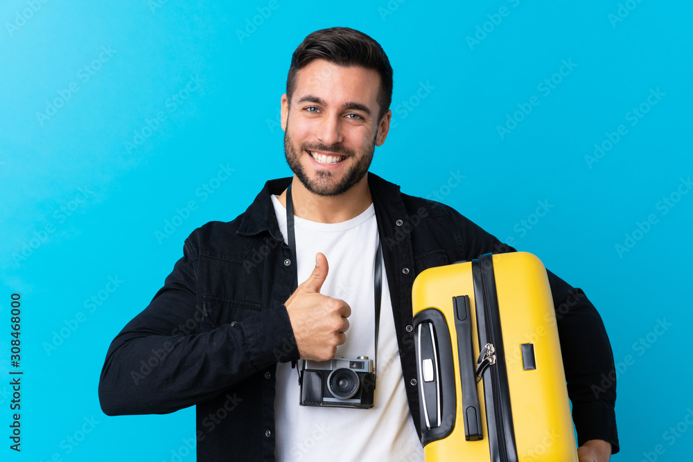 Traveler man holding a suitcase over isolated blue background with thumbs up because something good has happened Traveler man holding a suitcase over isolated blue background with thumbs up because something good has happened
