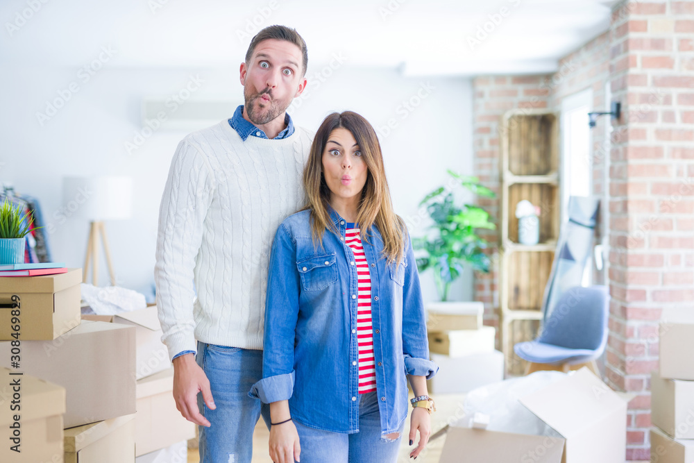 Obraz premium Young beautiful couple standing at new home around cardboard boxes making fish face with lips, crazy and comical gesture. Funny expression.