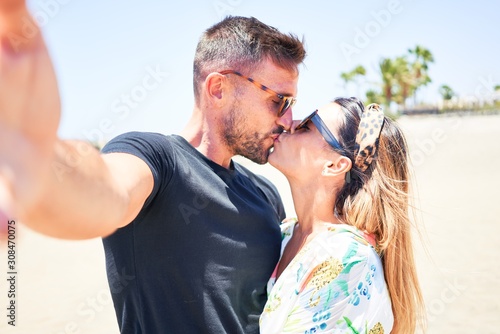 Young beautiful couple on vacation smiling happy and confident making selfie by camera. Standing with smile on face hugging and kissing at the beach