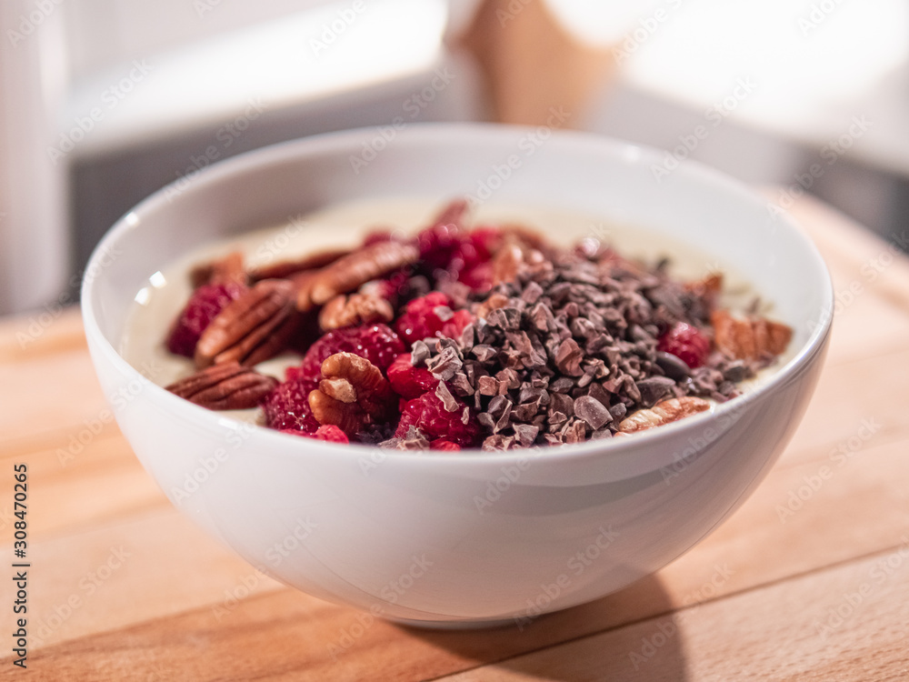 Eine weiße Keramikschüssel mit Joghurt, Pecannüssen, Kakaonibs und Himbeeren, steht in der Küche auf einem hellen Holzbrett. Eine weiße Keramikschüssel mit Joghurt, Pecannüssen, Kakaonibs und Himbeeren, steht in der Küche auf einem hellen Holzbrett.