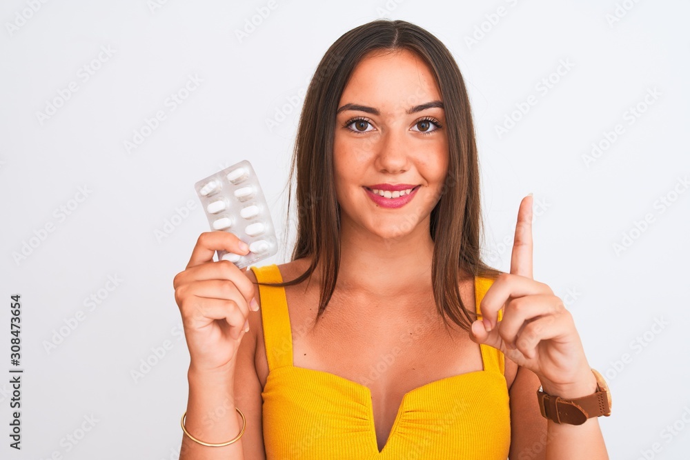 Young beautiful girl holding medicine pills standing over isolated white background surprised with an idea or question pointing finger with happy face, number one Young beautiful girl holding medicine pills standing over isolated white background surprised with an idea or question pointing finger with happy face, number one