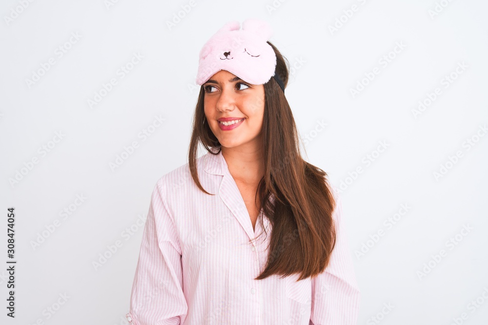 Young beautiful girl wearing pajama and sleep mask standing over isolated white background looking away to side with smile on face, natural expression. Laughing confident. Young beautiful girl wearing pajama and sleep mask standing over isolated white background looking away to side with smile on face, natural expression. Laughing confident.