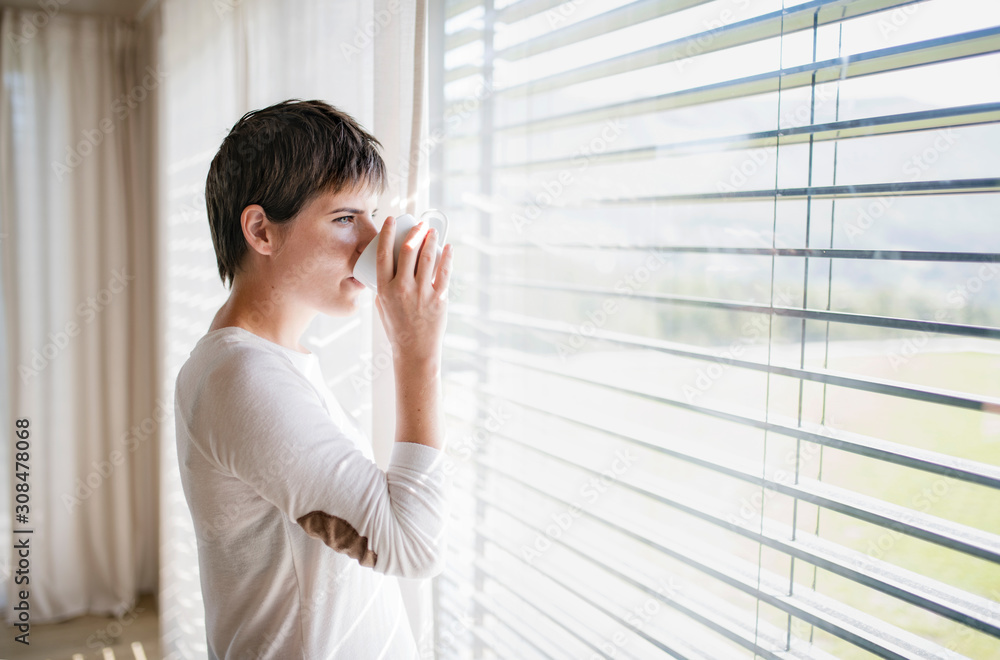 Portrait of young woman with coffee standing by window indoors at home. Portrait of young woman with coffee standing by window indoors at home.
