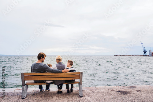 Rear view of young family with two small children on bench outdoors on beach.