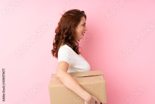 Young Russian woman over isolated pink background holding a box to move it to another site in lateral position © luismolinero